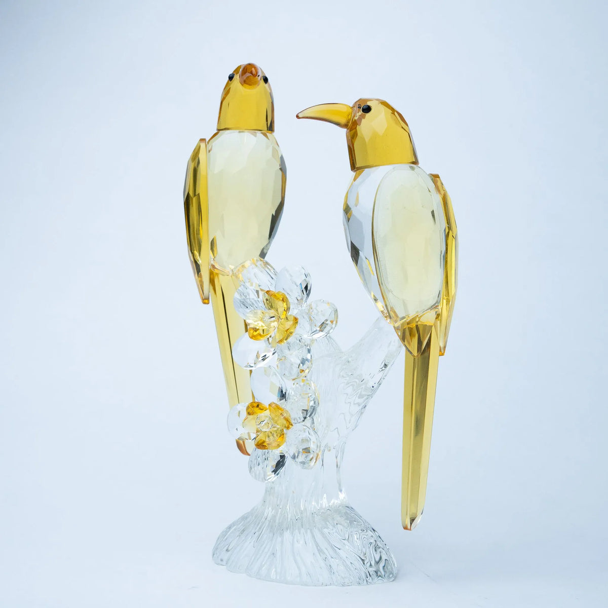 A pair of crystal bird figurines with translucent yellow bodies and clear wings, perched on a clear crystal branch with yellow flower accents