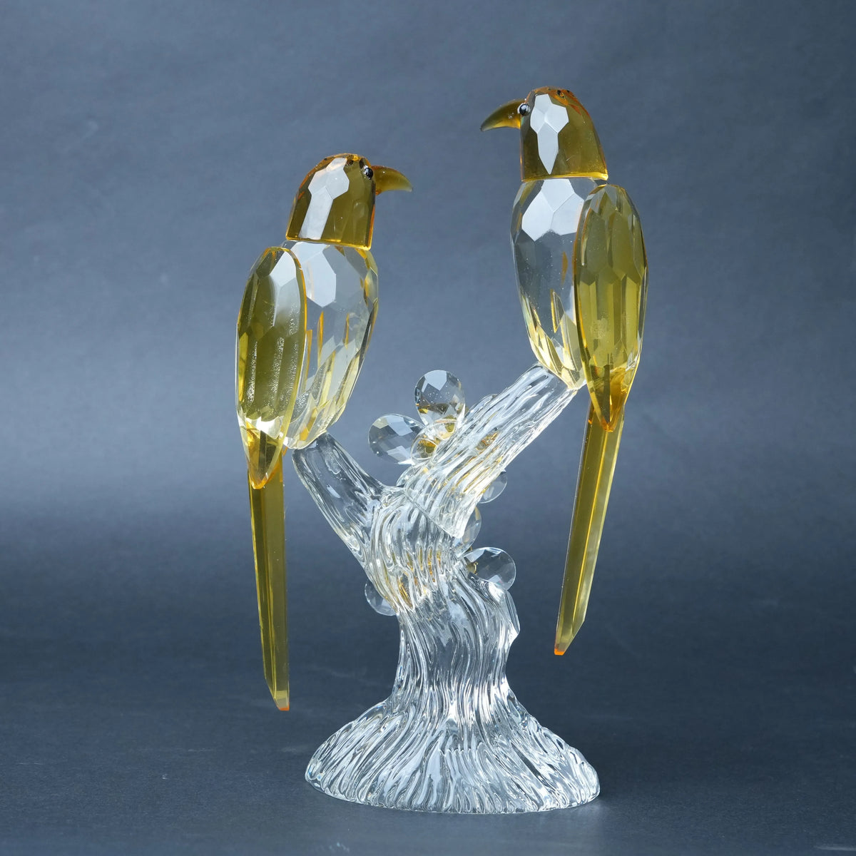 A decorative crystal bird sculpture featuring two faceted clear birds with translucent amber-yellow heads and tails, perched on a clear crystal branch base.