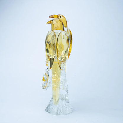 A decorative bird figurine made of faceted transparent and translucent materials, with a yellow-gold bird body and wings mounted on a clear crystal-like base.
