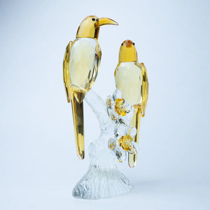 A decorative figurine of two golden birds perched on a clear glass branch with small yellow flowers