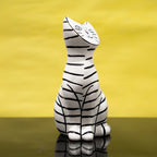 A resin cat figurine with a white base and black hand-drawn stripes, sitting upright on a reflective dark surface against a solid yellow background.