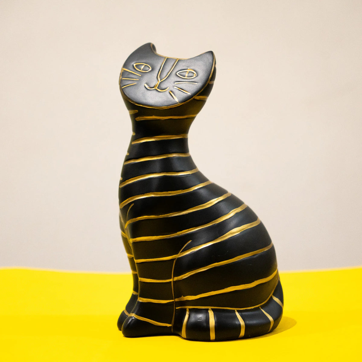 A stylized black cat figurine with gold stripes, sitting upright against a two-tone beige and yellow background.
