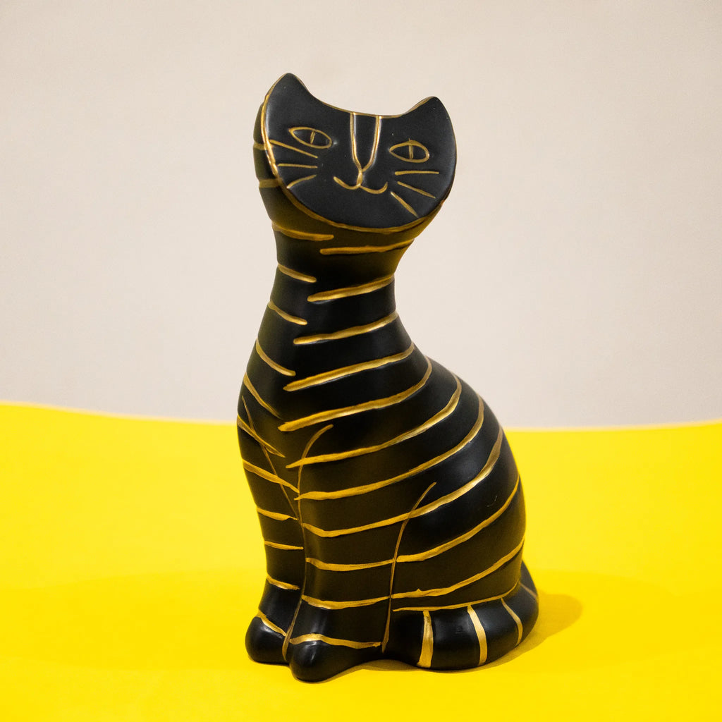 A black cat figurine with gold stripes, sitting upright on a bright yellow surface against a beige background.
