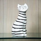 A ceramic cat figurine with a white body and black hand-drawn stripes, featuring a stylized face on its head, displayed on a glass shelf against a light wood-paneled background.