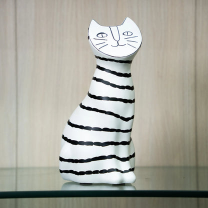 A ceramic cat figurine with a white body and black hand-drawn stripes, featuring a stylized face on its head, displayed on a glass shelf against a light wood-paneled background.