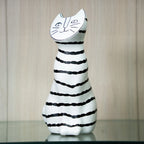 A ceramic cat-shaped candle holder with a white body, black horizontal stripes, and a white cat face on the top, displayed on a reflective surface against a light wood-paneled background.