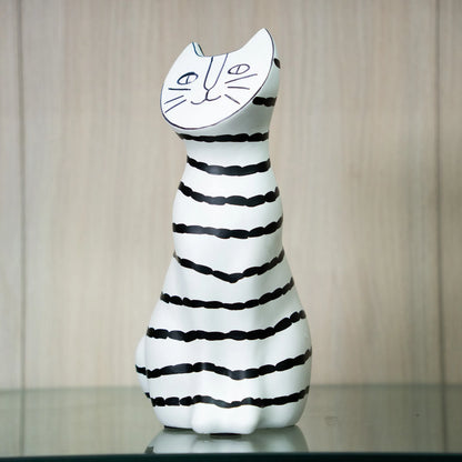A ceramic cat-shaped candle holder with a white body, black horizontal stripes, and a white cat face on the top, displayed on a reflective surface against a light wood-paneled background.