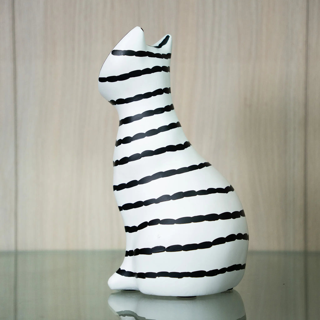 A white ceramic cat figurine with black hand-drawn style horizontal stripes, sitting on a reflective glass surface against a light wood-paneled background.