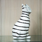 A white ceramic cat figurine with hand-drawn black stripes and a simple black face, sitting on a reflective surface against a light wood-paneled background.
