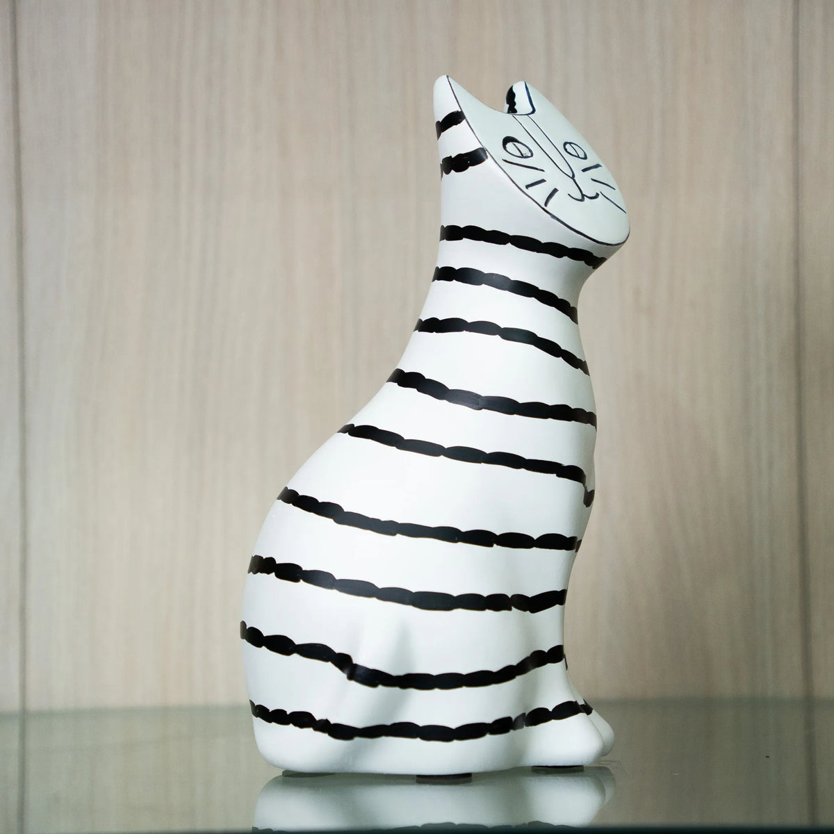 A white ceramic cat figurine with hand-drawn black stripes and a simple black face, sitting on a reflective surface against a light wood-paneled background.