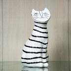 A ceramic cat figurine with a white body and black horizontal stripes, featuring a stylized cat face on its head, displayed on a reflective surface against a light wood background.