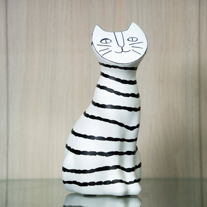 A ceramic cat figurine with a white body and black horizontal stripes, featuring a stylized cat face on its head, displayed on a reflective surface against a light wood background.
