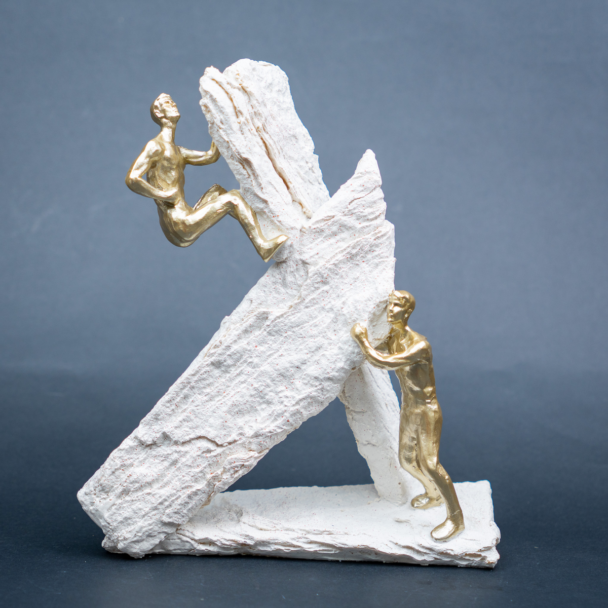 A sculptural figurine depicting two gold human figures against a large white textured rock formation, photographed on a dark gray studio background.
