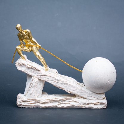 A small decorative sculpture showing a gold human figure pulling a large white sphere with a string, mounted on an off-white textured base, photographed against a dark gray studio background.