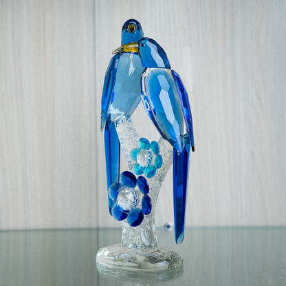 A blue and clear glass bird figurine perched on a transparent glass stand, with small flower details and a yellow beak accent, photographed against a light wood background.