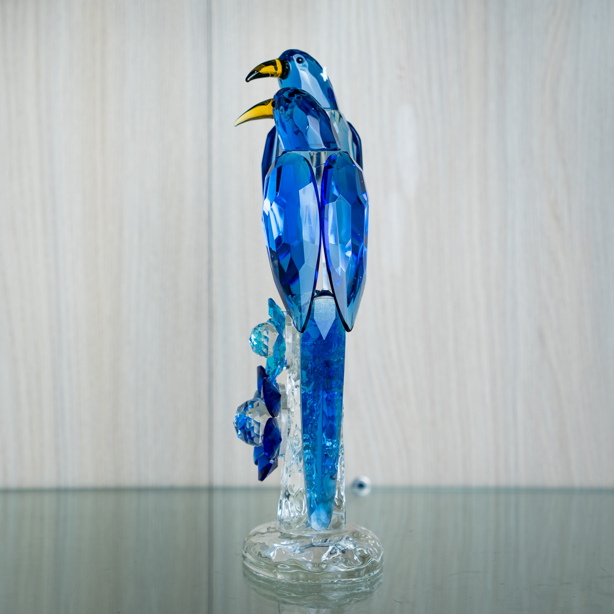 A blue faceted crystal bird sculpture perched on a clear glass base, photographed against a light wood-paneled wall and reflective glass surface.