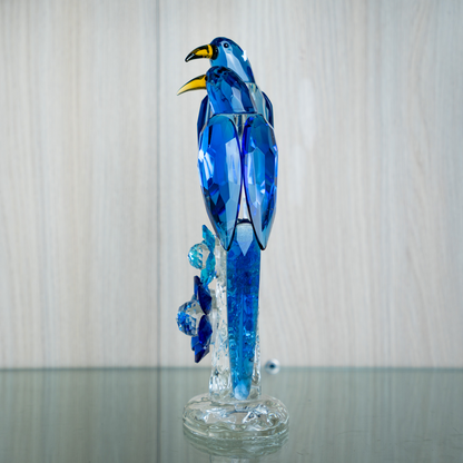 A blue faceted crystal bird sculpture perched on a clear glass base, photographed against a light wood-paneled wall and reflective glass surface.