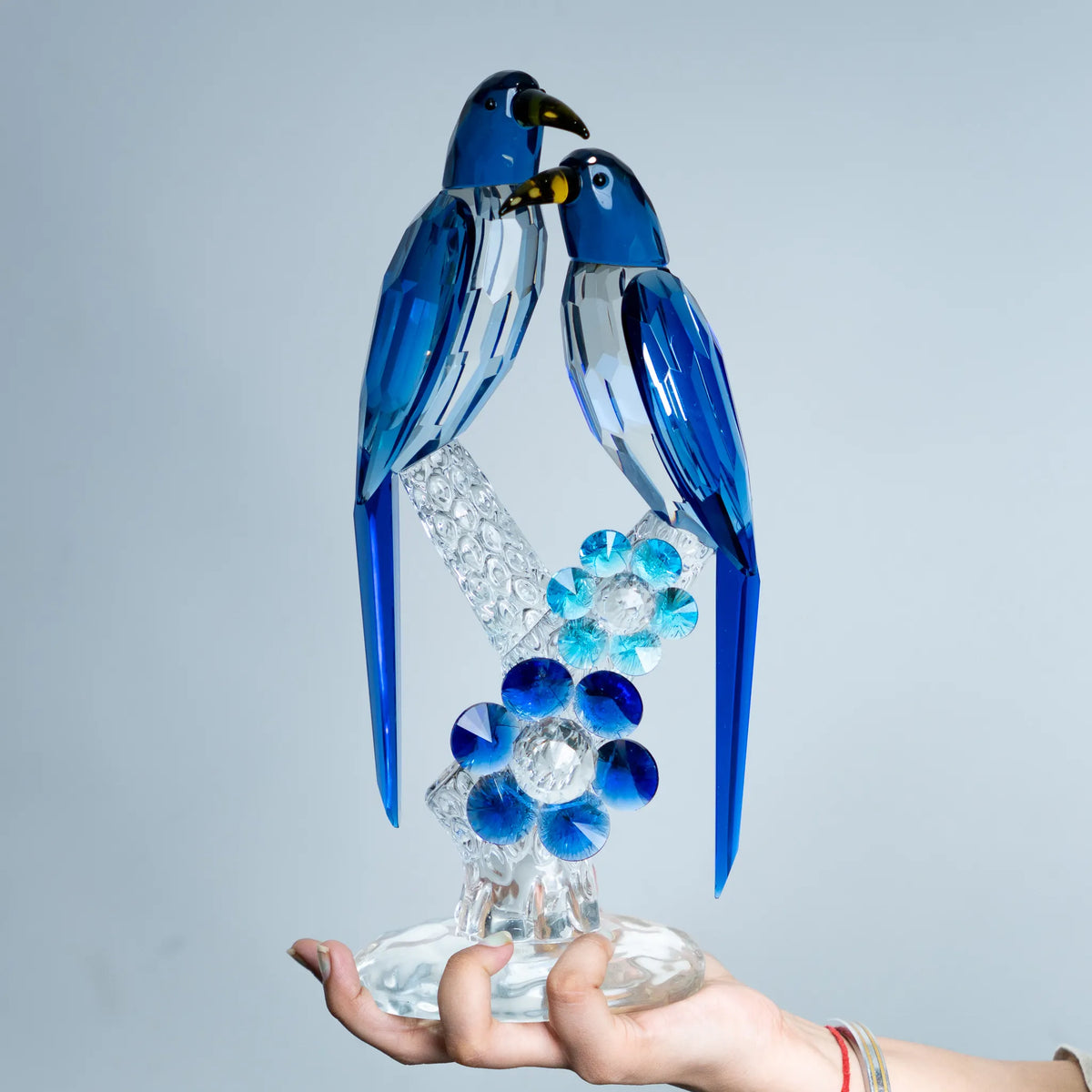 A hand holds a blue and clear crystal bird sculpture of two birds perched on a flower, against a plain light gray background.
