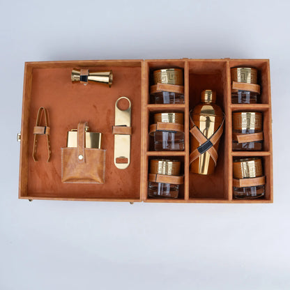 A luxury bar gift set in a wooden presentation box, featuring a gold-toned bottle with a leather wrap, six small amber glass jars with gold lids, a leather pouch with gold accessories, and a brass tongs, all arranged neatly inside an open light brown leather.