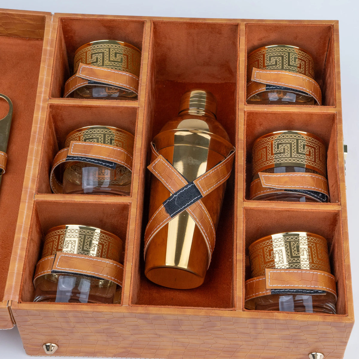 A wooden gift box with six compartments holding four clear glass tumblers with gold Greek key bands and brown leather straps, plus a central gold bottle wrapped with a brown leather strap.