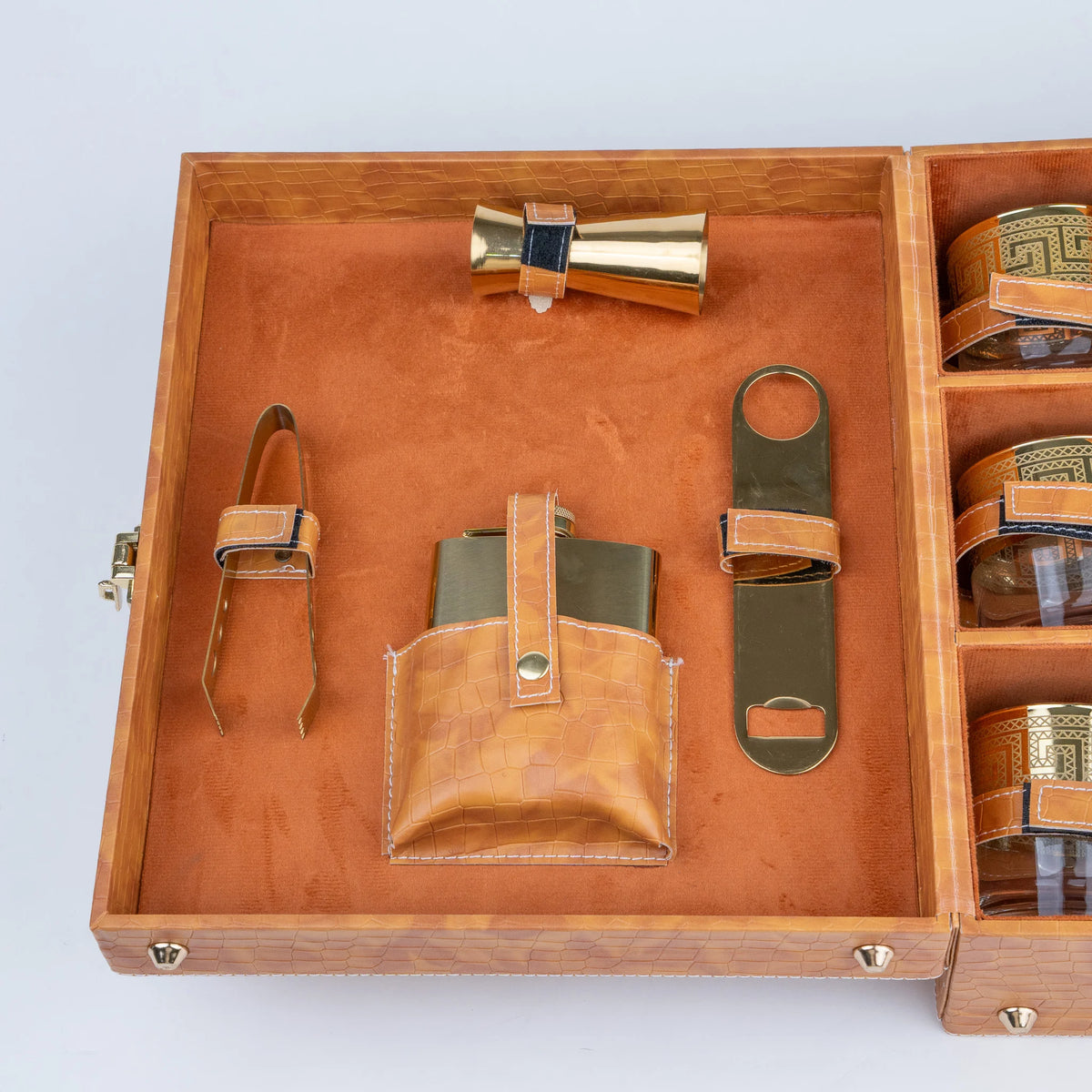 An open wooden presentation box containing a leather flask pouch, bottle opener, bottle tongs, and a small gold-toned accessory, with additional compartments holding round glass containers.