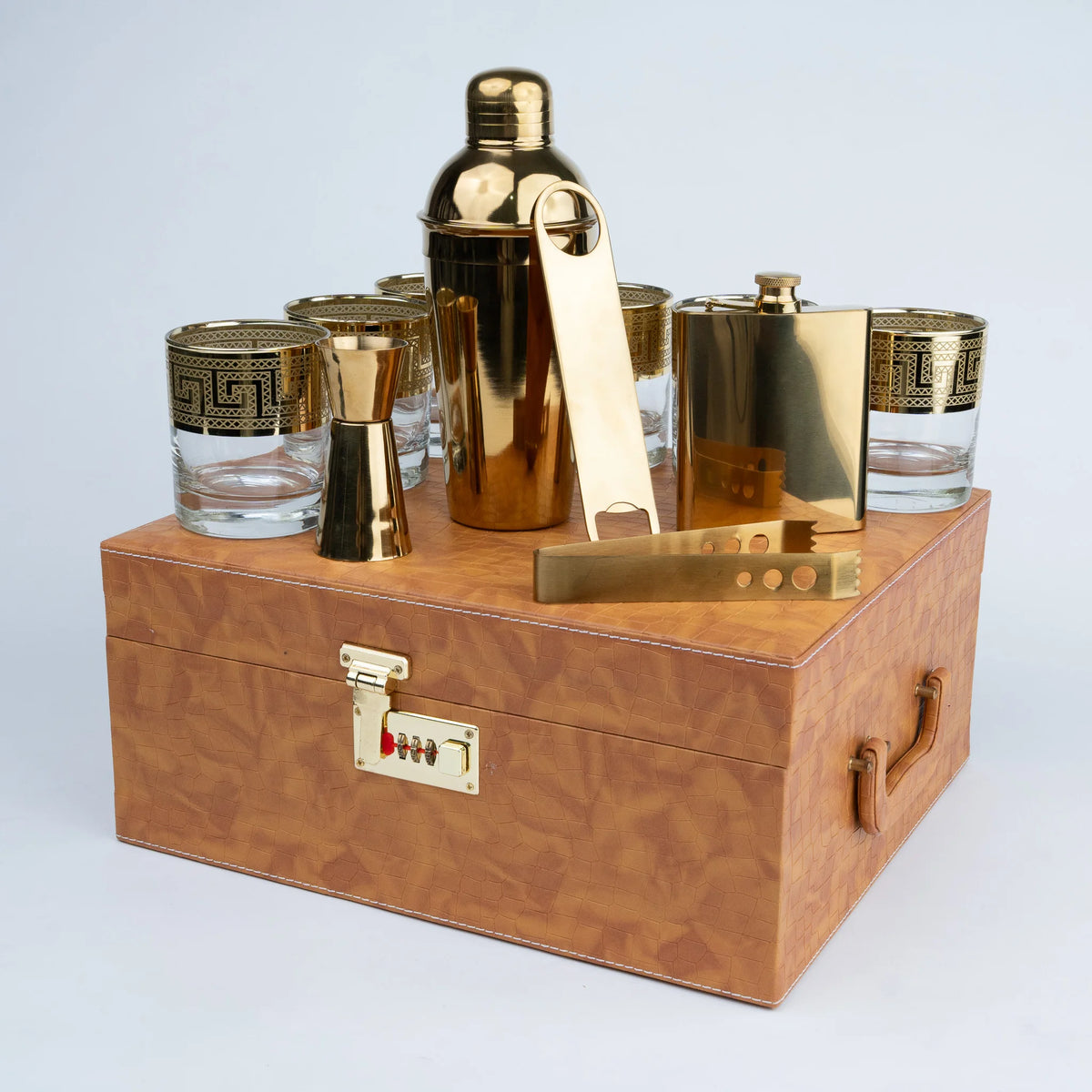 A luxury bar gift set in a wooden box, featuring a gold shaker, flask, jigger, and tumblers with gold Greek key patterns,
