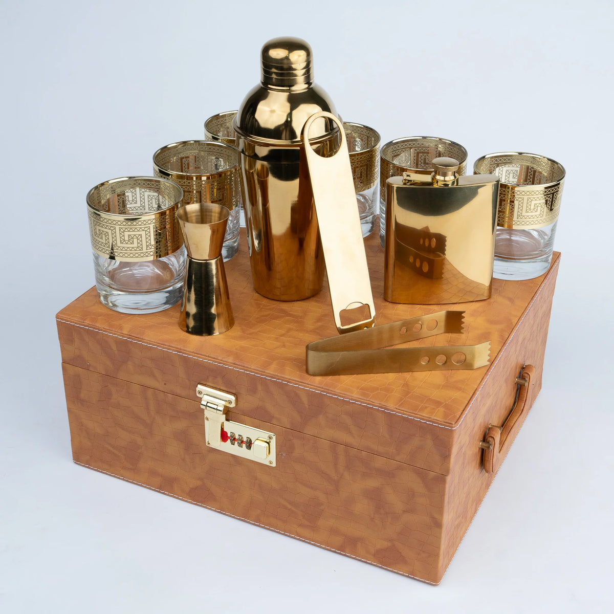 A gold and brown cocktail gift set displayed on a matching wooden case The set includes a gold shaker, several gold and clear glasses, a jigger, a flask, and tongs.