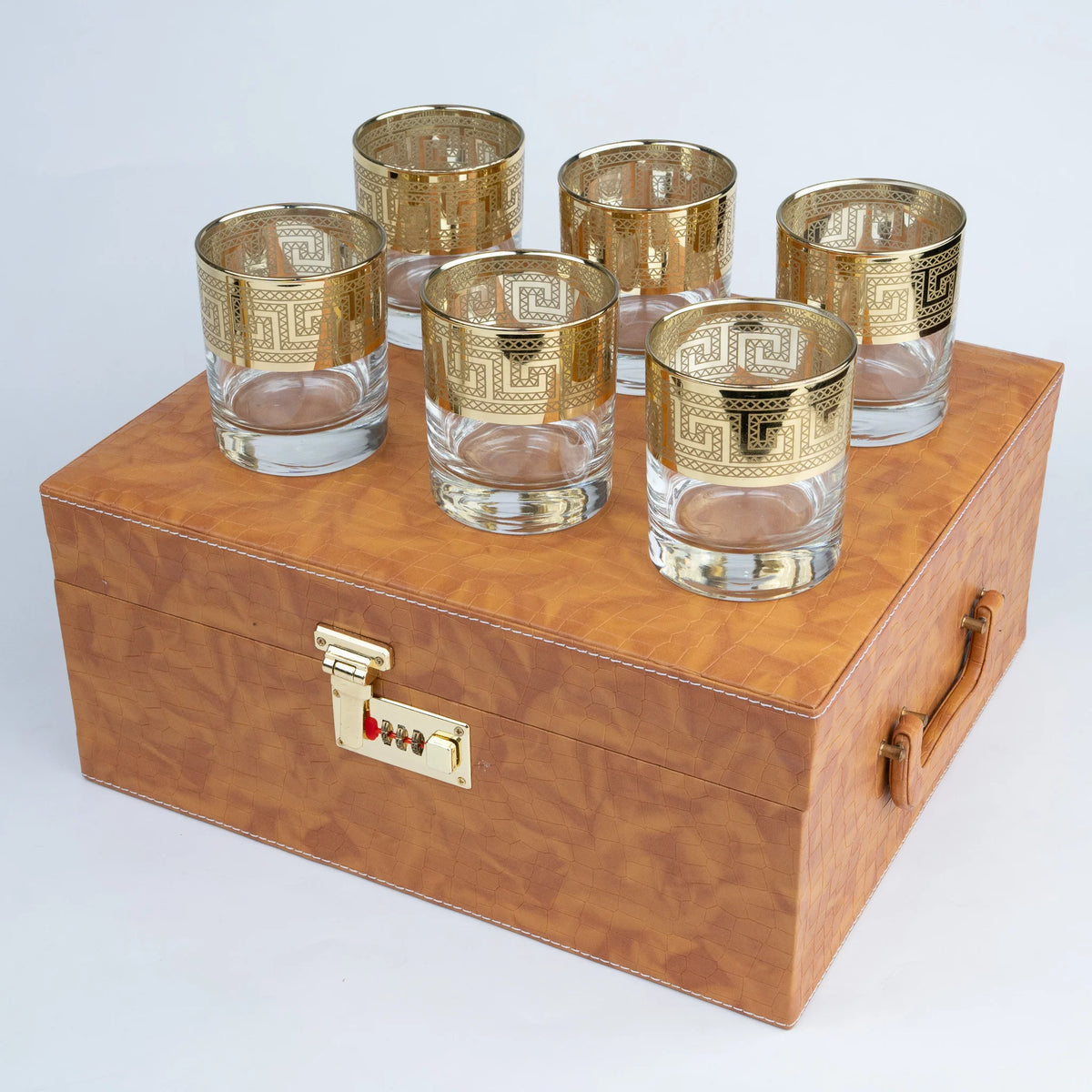 A set of six gold-patterned glass tumblers arranged on top of a brown wood-grain storage box with a gold combination lock and handle