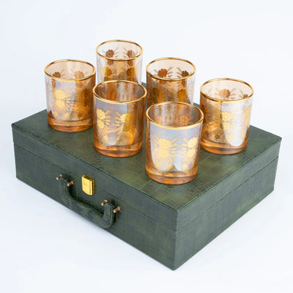 Six amber-tinted glass tumblers with gold floral etched patterns, arranged on top of a dark green textured presentation box with a gold clasp, against a white background.