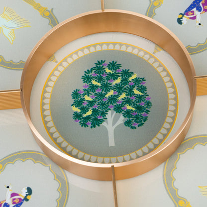 A circular copper-toned metal tray or dish featuring a printed illustration of a green tree with purple and yellow leaves on a pale gray background, surrounded by a scalloped gold border.