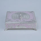 A decorative silver jewelry box with a light pink enamel or lacquer base, featuring embossed peacock and floral motifs, ornate silver scrollwork borders, and small silver feet.