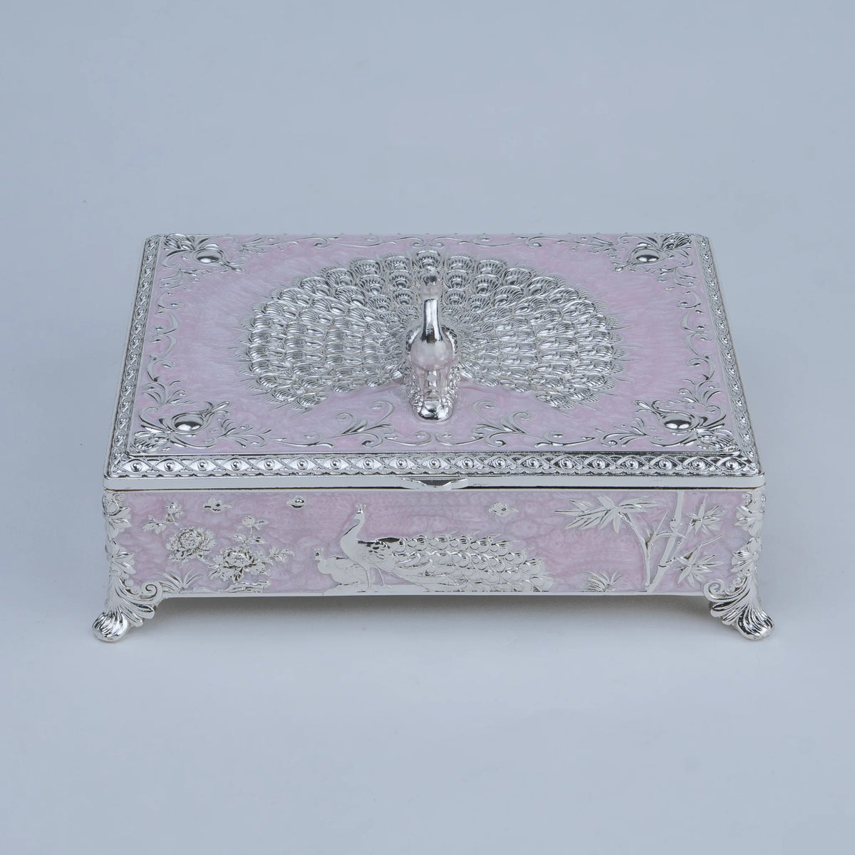 A decorative silver jewelry box with a light pink enamel or lacquer base, featuring embossed peacock and floral motifs, ornate silver scrollwork borders, and small silver feet.