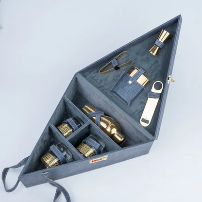 A dark gray wooden cocktail gift set organizer shown open, containing gold-toned metal bar tools and bottles with dark blue leather straps.