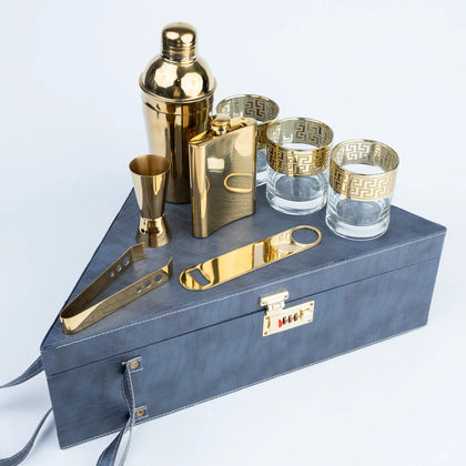 A gold-toned cocktail bar gift set arranged on a dark blue textured box, including a shaker, jigger, flask, bottle opener, tongs, and patterned glasses.