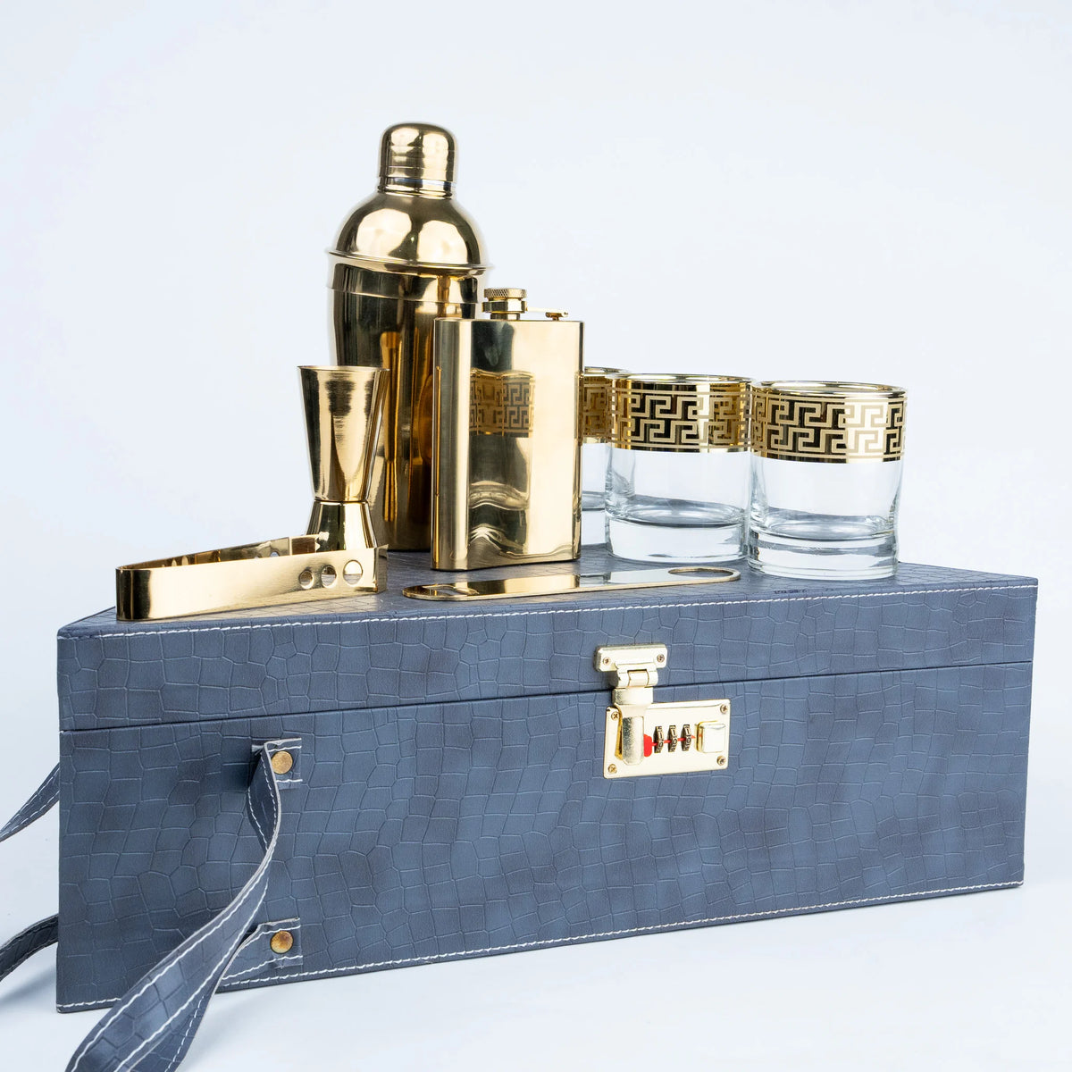 A luxury bar gift set featuring a gold shaker, flask, jigger, and two glasses with Greek key detailing, displayed on a blue textured leather case with a lock and carrying strap.