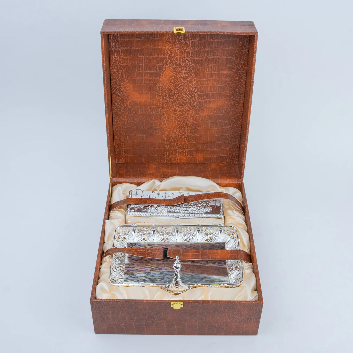 An open wooden presentation box with a crocodile-embossed interior lid and cream satin lining, holding three rectangular silver trays with decorative cutout patterns and small hanging silver charms, photographed on a light gray studio background.