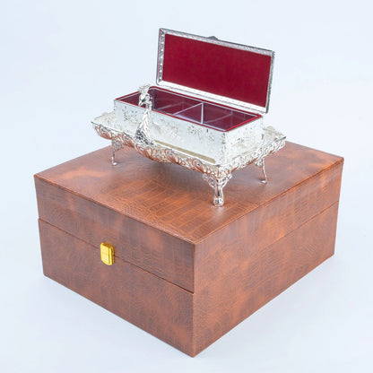 A silver-toned ornate jewelry box with a red velvet-lined interior sits on top of a brown textured wooden storage box with a gold clasp, photographed on a light gray studio background.