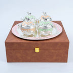 A set of four decorative sugar bowls with silver lids and pastel-colored interiors, presented on a round silver tray atop a brown box with a gold clasp.