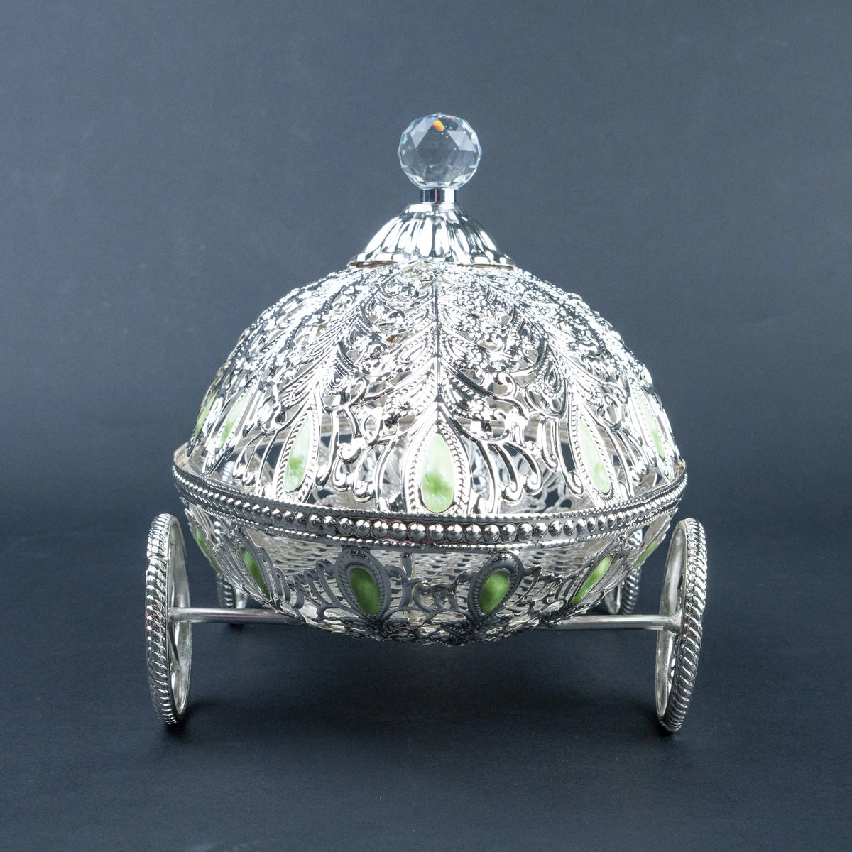 An ornate silver-toned decorative box shaped like a carriage, with intricate filigree detailing, small green stone accents, and a clear faceted crystal knob on top