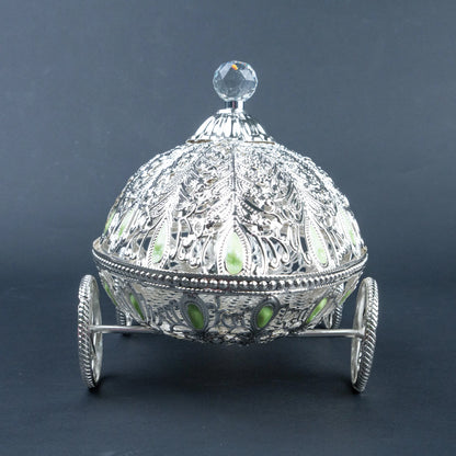 An ornate silver-toned decorative box shaped like a carriage, with intricate filigree detailing, small green stone accents, and a clear faceted crystal knob on top