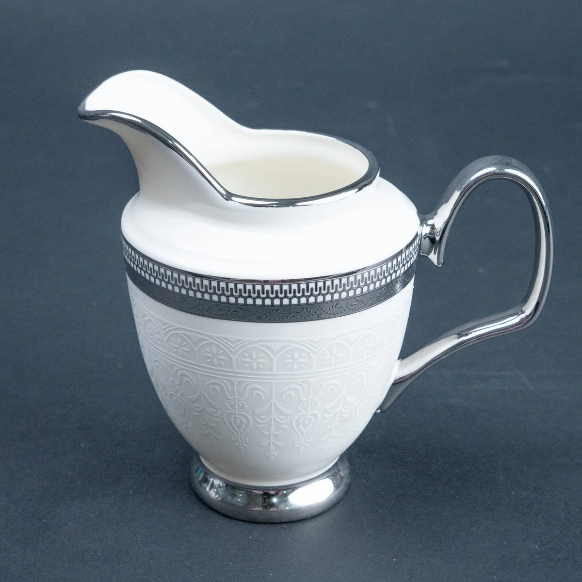 A white ceramic creamer pitcher with embossed floral detailing, a silver rim, and a polished silver handle, set against a dark gray background.