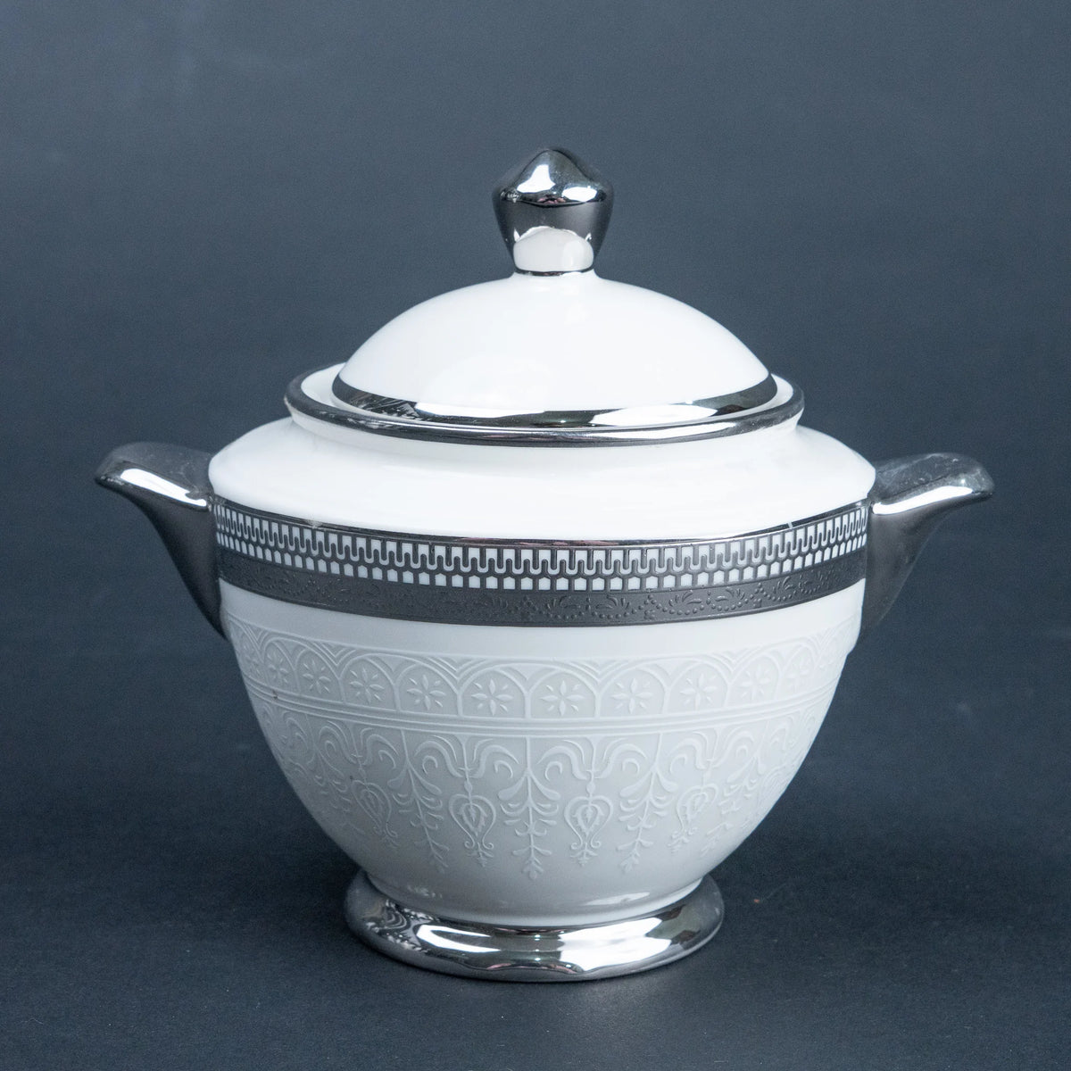 A white ceramic lidded sugar bowl or creamer with silver-toned metal handles, spout, lid knob, and decorative bands, photographed on a dark gray studio background.