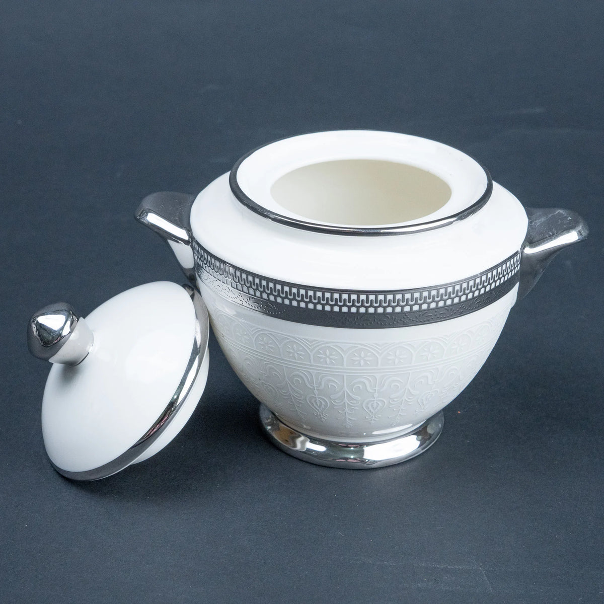 A white ceramic sugar bowl with a matching lid, featuring silver-tone metal handles, rim detailing, and a decorative embossed pattern on the body, set against a dark gray background.
