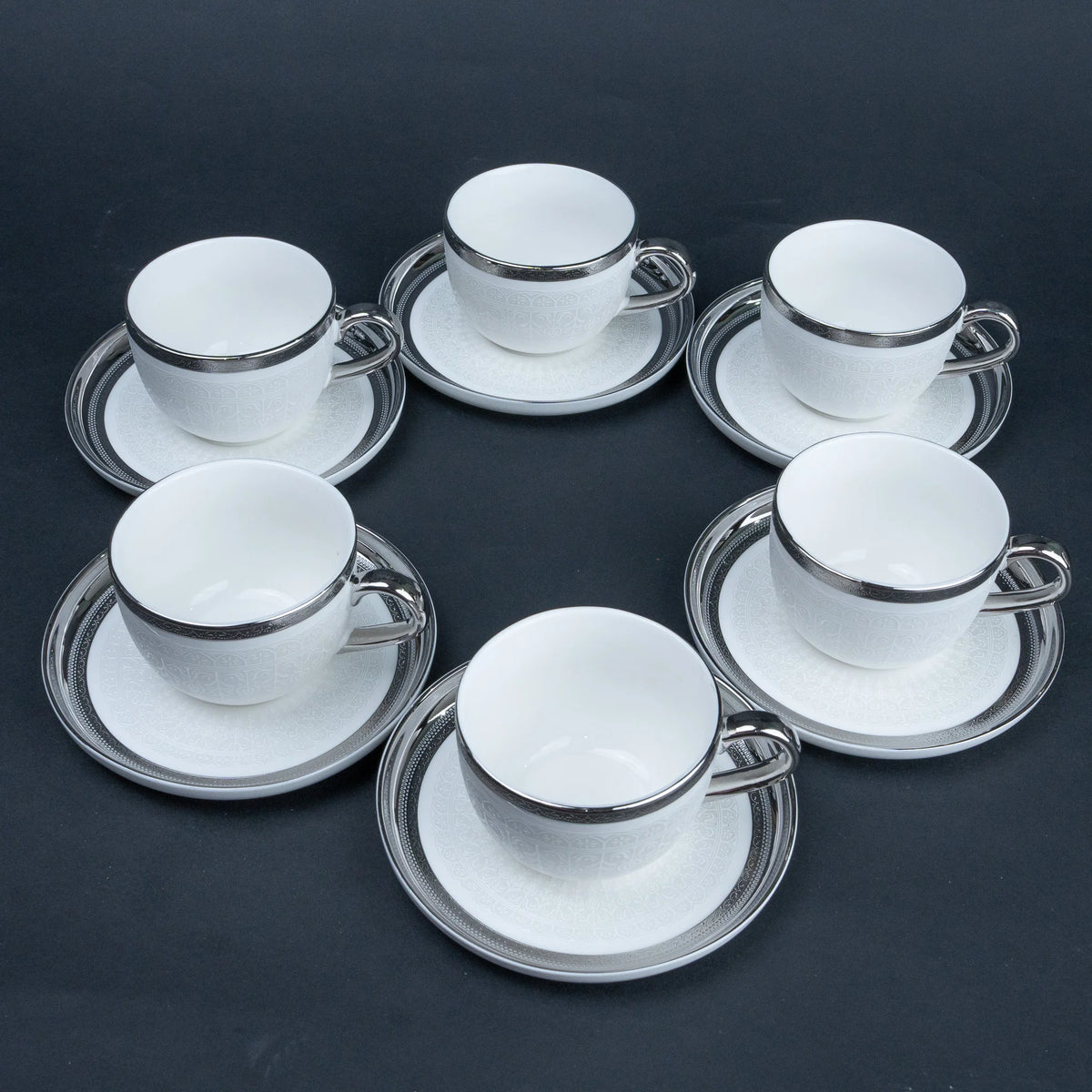 Six white ceramic espresso cups with matching saucers, each accented with a polished silver rim, arranged in a circular pattern on a dark gray background.