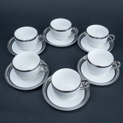 Six white ceramic espresso cups with matching saucers, each accented with a polished silver rim, arranged in a circular pattern on a dark gray background.