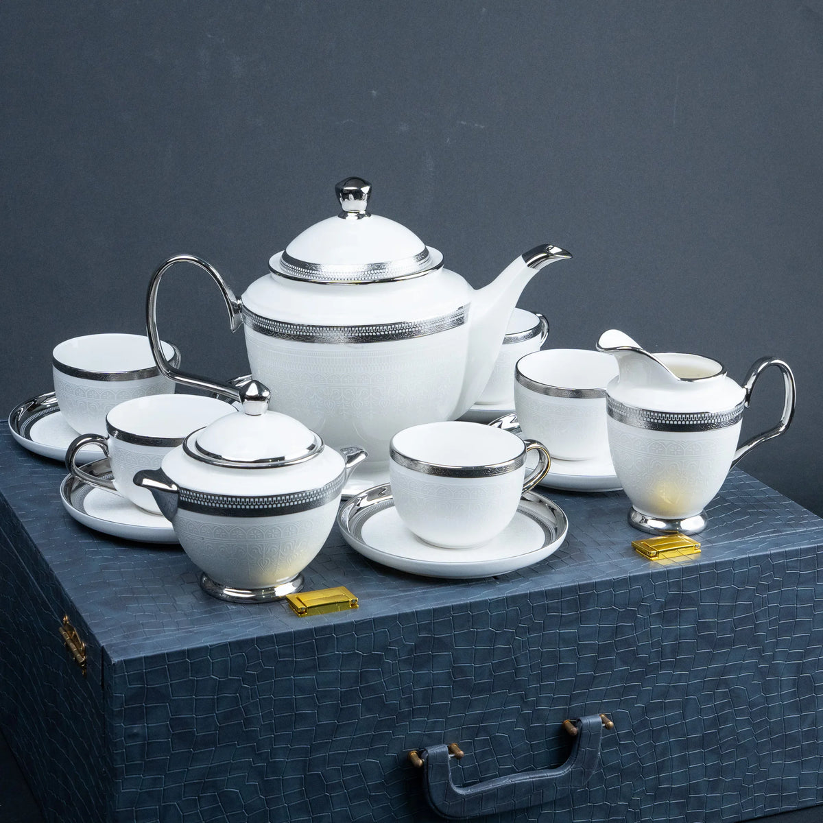 A white porcelain tea and coffee set with silver trim, including a teapot, teacups, sugar bowl, creamer, and small containers, arranged on a dark textured surface against a dark gray background.