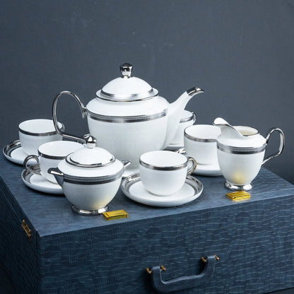 A white porcelain tea and coffee set with silver trim, including a teapot, teacups, sugar bowl, creamer, and small containers, arranged on a dark textured surface against a dark gray background.