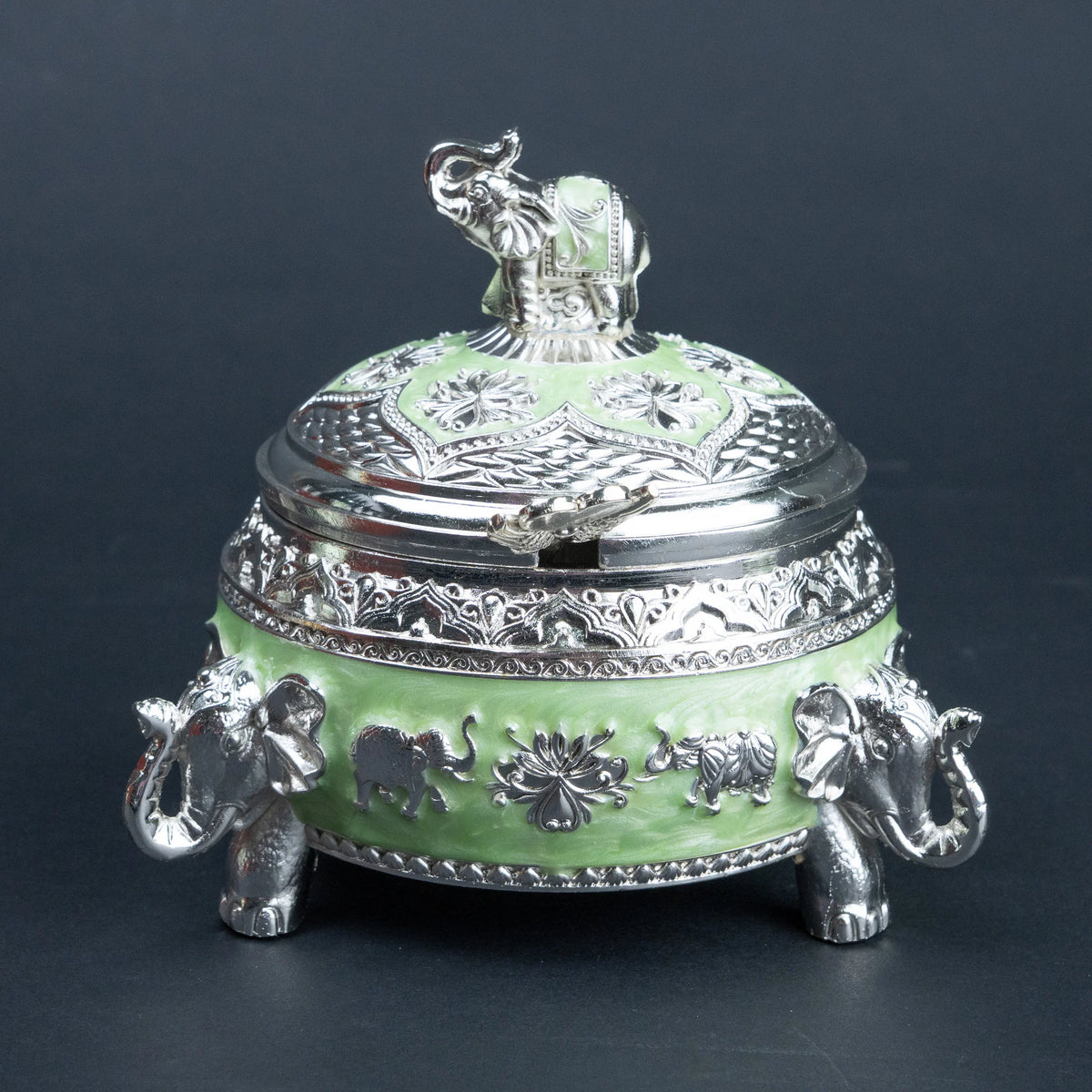 A decorative lidded box with a light green enamel body and ornate silver-tone metal detailing, including elephant figures on the base and lid, photographed against a dark gray background.