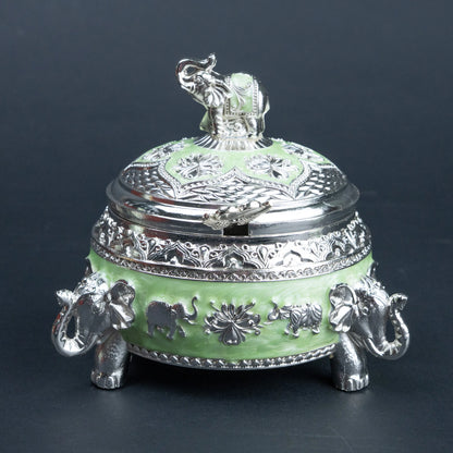 A decorative lidded box with a light green enamel body and ornate silver-tone metal detailing, including elephant figures on the base and lid, photographed against a dark gray background.