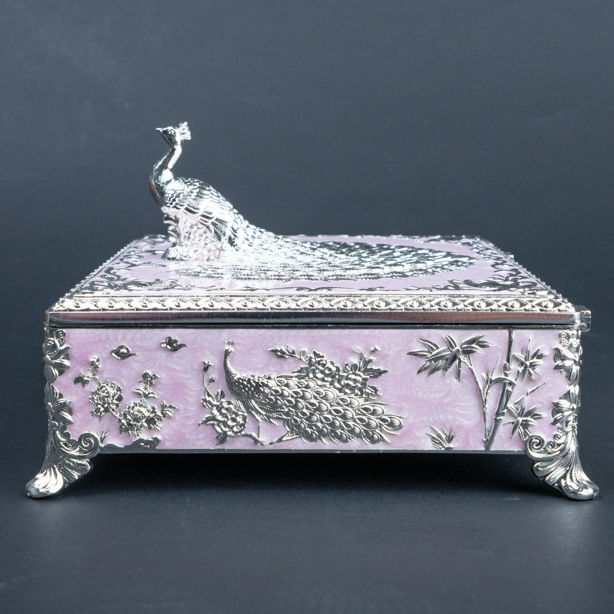A decorative rectangular box with a lavender-pink enamel-like finish and raised silver-tone peacock and floral motifs, topped with a sculpted peacock figure on a dark gray background.